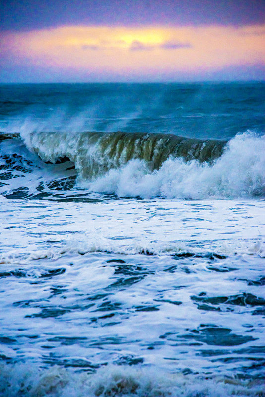 Large wave breaking against the shore at sunset with purple and yellow sky behind
